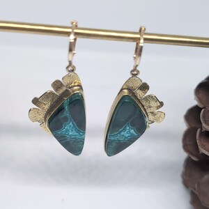 May include: A pair of dangle earrings featuring triangular dark green stones with intricate teal veining. Each stone is set in a textured gold-tone metal frame with a fan-like decorative edge. The earrings hang from small gold-tone hoop closures.