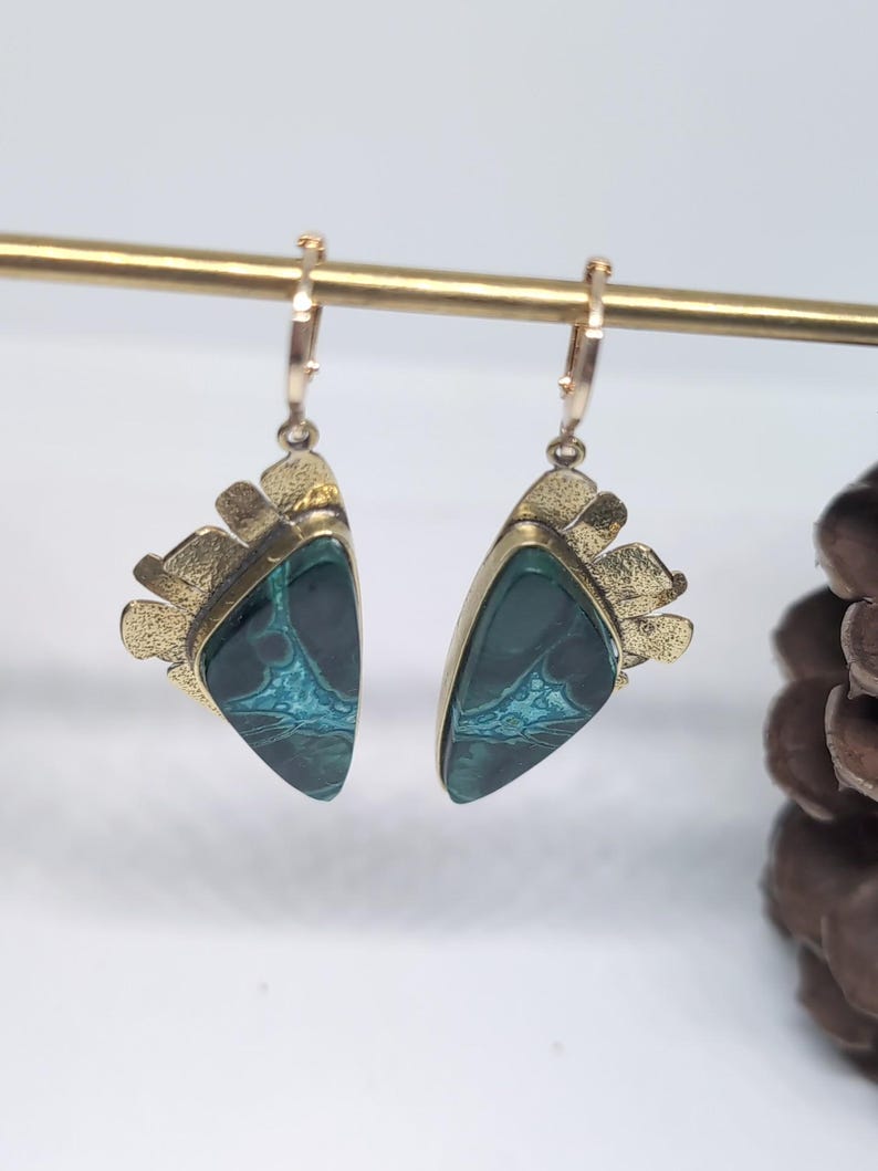 May include: A pair of dangle earrings featuring triangular dark green stones with intricate teal veining. Each stone is set in a textured gold-tone metal frame with a fan-like decorative edge. The earrings hang from small gold-tone hoop closures.