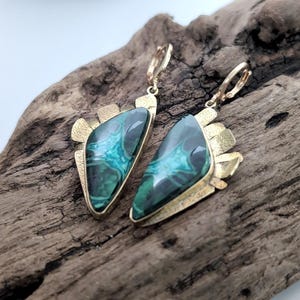 May include: A pair of dangle earrings featuring triangular malachite stones with deep green and teal banding. The stones are set in textured gold-tone metal frames with a jagged, fan-like edge and secure lever-back closures for attachment.