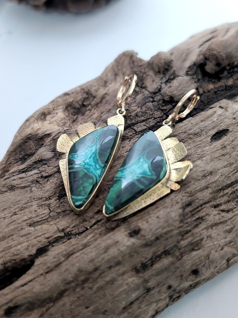 May include: A pair of dangle earrings featuring triangular malachite stones with deep green and teal banding. The stones are set in textured gold-tone metal frames with a jagged, fan-like edge and secure lever-back closures for attachment.