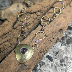 May include: A necklace featuring a gold-toned triangular pendant with a central purple stone, connected to a chain of alternating gold-toned circular links and small purple beads. The metal surface of the pendant has a textured, hammered finish.
