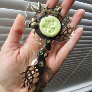May include: A decorative metal brooch featuring a central circular pale green cabochon with an embossed frog design. The piece has an oxidized dark metal finish with polished gold-tone accents, leaf motifs, and three small green inset stones.