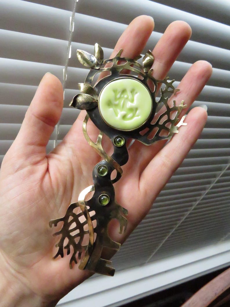 May include: A decorative metal brooch featuring a central circular pale green cabochon with an embossed frog design. The piece has an oxidized dark metal finish with polished gold-tone accents, leaf motifs, and three small green inset stones.