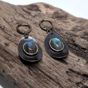 May include: A pair of drop earrings featuring oval labradorite gemstones with blue iridescence, set in dark bronze-toned metal bezels. The earrings have a teardrop-shaped backing and are attached to circular lever-back clasps in a matching bronze finish.