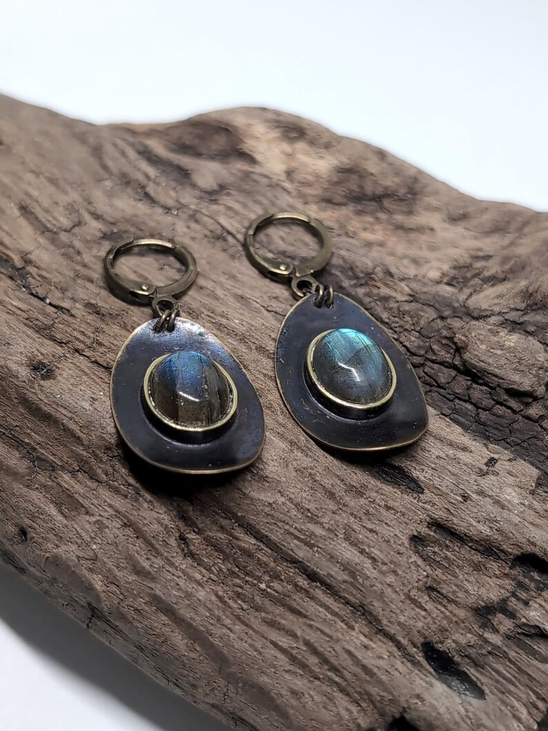 May include: A pair of drop earrings featuring oval labradorite gemstones with blue iridescence, set in dark bronze-toned metal bezels. The earrings have a teardrop-shaped backing and are attached to circular lever-back clasps in a matching bronze finish.