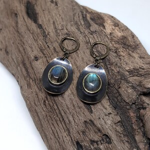 May include: A pair of drop earrings featuring oval-shaped labradorite gemstones with blue iridescence, set in dark, oxidized metal frames. The earrings are attached to circular lever-back clasps made of a matching antique bronze-toned metal finish.