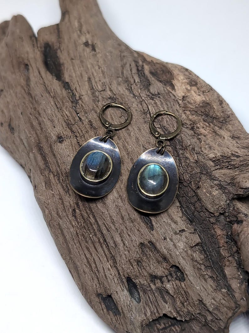 May include: A pair of drop earrings featuring oval-shaped labradorite gemstones with blue iridescence, set in dark, oxidized metal frames. The earrings are attached to circular lever-back clasps made of a matching antique bronze-toned metal finish.