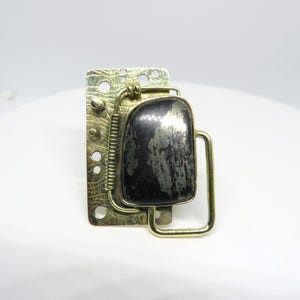 Puede incluir: Broche artesanal con una piedra rectangular gris oscuro engastada en un marco de latón. El latón tiene un diseño industrial texturizado con un resorte y recortes circulares. Un broche llamativo.