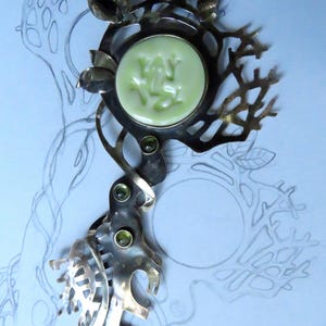 May include: A silver-toned metal brooch featuring an intricate, branch-like openwork design. The piece is centered with a circular, pale green cabochon embossed with a frog figure and accented by three small, round, dark green inset stones.