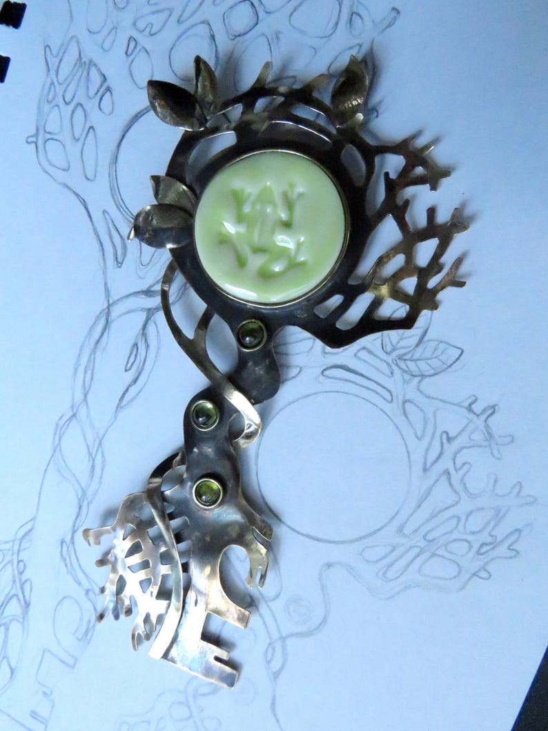 May include: A silver-toned metal brooch featuring an intricate, branch-like openwork design. The piece is centered with a circular, pale green cabochon embossed with a frog figure and accented by three small, round, dark green inset stones.