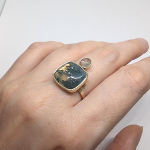 May include: A gold ring featuring a large, square-shaped moss agate stone with dark, dendritic inclusions and a smaller, round, translucent stone set beside it. The band is thin and metallic, showcasing a polished gold finish throughout the setting.