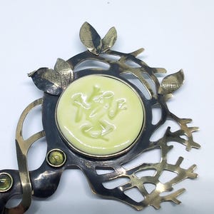 May include: A decorative metal brooch featuring a central pale yellow circular ceramic inlay with a relief of a frog. The metal frame is dark with a branch-like structure, accented by small metallic leaves and two small green circular stones.