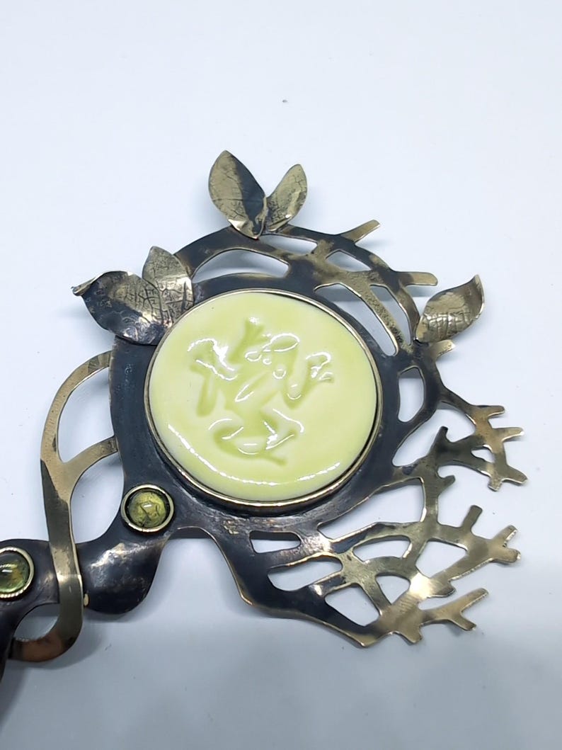 May include: A decorative metal brooch featuring a central pale yellow circular ceramic inlay with a relief of a frog. The metal frame is dark with a branch-like structure, accented by small metallic leaves and two small green circular stones.