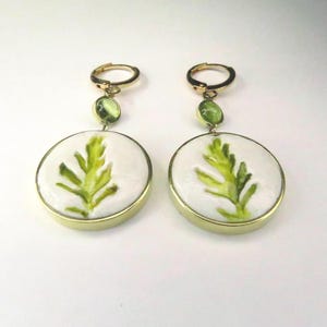 May include: A pair of gold-toned dangle earrings featuring circular white ceramic discs with a painted green leaf design. Each earring includes a small, round, light green gemstone accent set above the disc, attached to a gold-toned hoop closure.