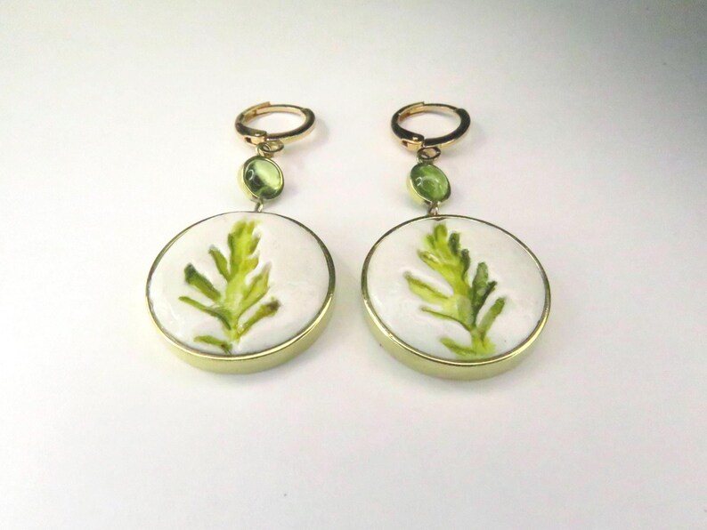 May include: A pair of gold-toned dangle earrings featuring circular white ceramic discs with a painted green leaf design. Each earring includes a small, round, light green gemstone accent set above the disc, attached to a gold-toned hoop closure.