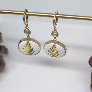 May include: A pair of gold-toned drop earrings featuring a circular white pendant with a green leaf illustration. Each earring includes a small, round, light green gemstone accent positioned above the circular pendant on a gold-toned hoop wire.