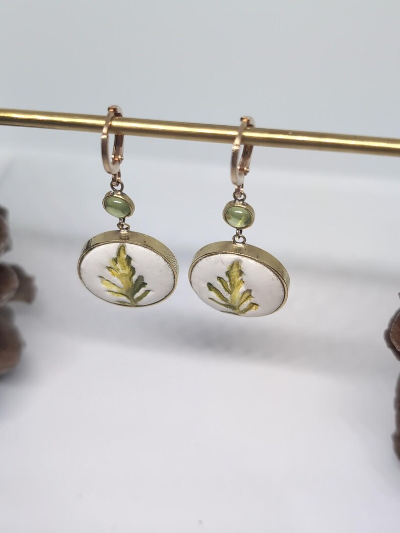 May include: A pair of gold-toned drop earrings featuring a circular white pendant with a green leaf illustration. Each earring includes a small, round, light green gemstone accent positioned above the circular pendant on a gold-toned hoop wire.
