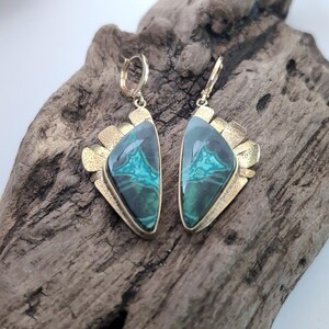 May include: A pair of drop earrings featuring triangular-shaped green and black malachite stones set in textured gold-tone metal frames. Each earring has a hinged hoop closure at the top and a decorative fan-like metal detail along the outer edge.