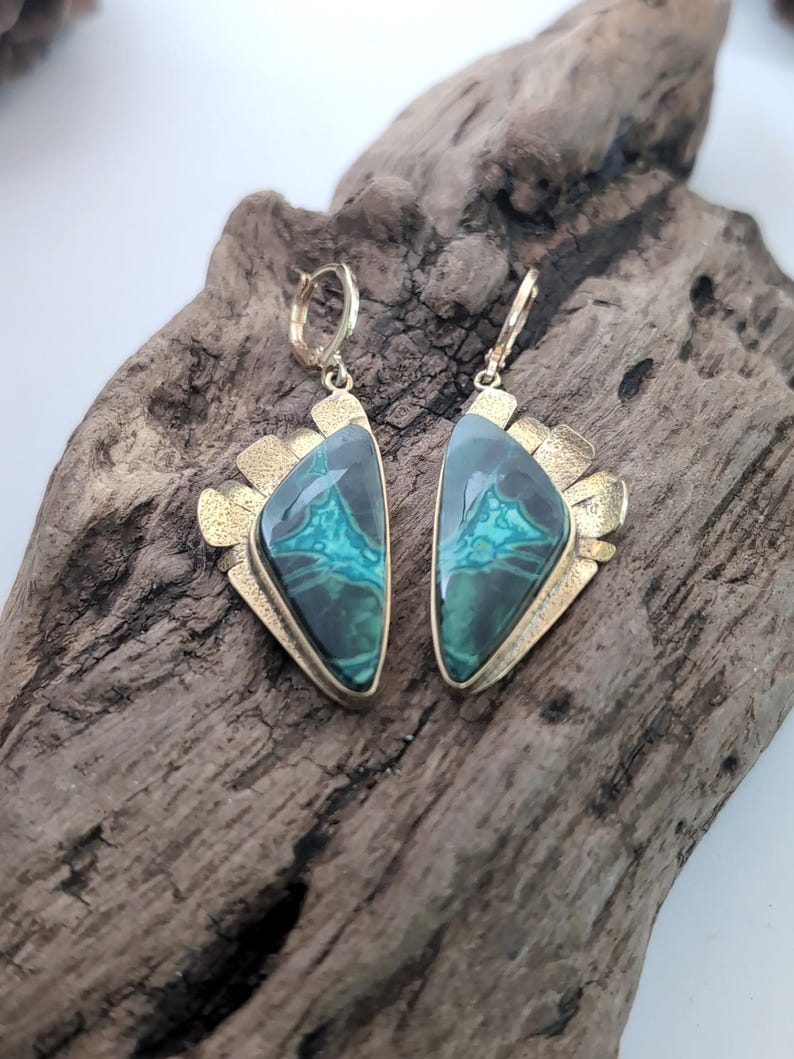 May include: A pair of drop earrings featuring triangular-shaped green and black malachite stones set in textured gold-tone metal frames. Each earring has a hinged hoop closure at the top and a decorative fan-like metal detail along the outer edge.