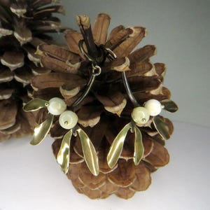 May include: A pair of gold-tone earrings with a mistletoe design. The earrings feature white faux pearls and are set against a brown pine cone background.