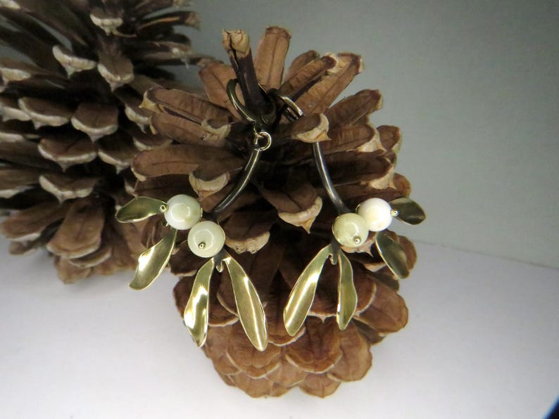 May include: A pair of gold-tone earrings with a mistletoe design. The earrings feature white faux pearls and are set against a brown pine cone background.