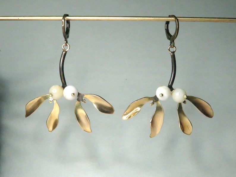 May include: A pair of gold mistletoe earrings with white pearl accents. The earrings are suspended from black metal hooks.