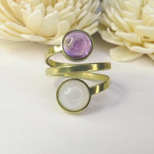 May include: A bypass ring crafted from gold-toned metal featuring two circular settings. The top setting holds a smooth, translucent purple cabochon, while the bottom setting contains a solid, opaque white cabochon. The band wraps around the finger.