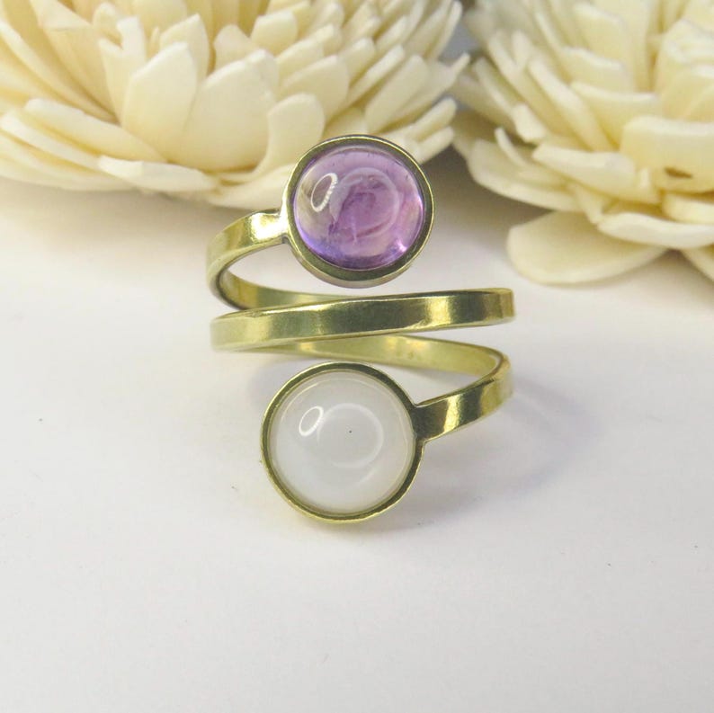 May include: A bypass ring crafted from gold-toned metal featuring two circular settings. The top setting holds a smooth, translucent purple cabochon, while the bottom setting contains a solid, opaque white cabochon. The band wraps around the finger.