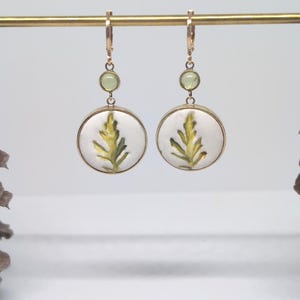 May include: A pair of gold-toned drop earrings featuring a circular white pendant with a green fern leaf illustration. Each earring includes a small, round, light green accent stone positioned above the main circular pendant on a gold wire hook.