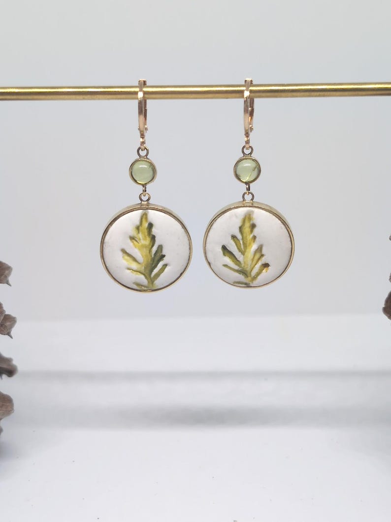 May include: A pair of gold-toned drop earrings featuring a circular white pendant with a green fern leaf illustration. Each earring includes a small, round, light green accent stone positioned above the main circular pendant on a gold wire hook.