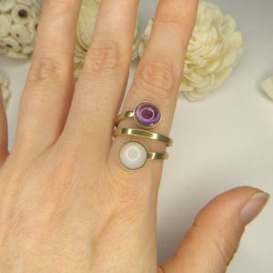May include: A gold-toned bypass ring featuring two circular settings. The top setting contains a translucent purple stone, while the bottom setting holds a smooth, opaque white stone. The band wraps around the finger in a spiral design.