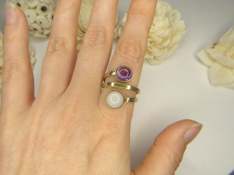 May include: A gold-toned bypass ring featuring two circular settings. The top setting contains a translucent purple stone, while the bottom setting holds a smooth, opaque white stone. The band wraps around the finger in a spiral design.