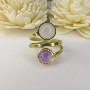May include: A bypass ring featuring a gold-toned metal band that spirals around the finger. The ring is adorned with two circular cabochon stones: one opaque white stone at the top and one translucent light purple stone at the bottom.