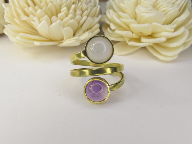 May include: A bypass ring featuring a gold-toned metal band that spirals around the finger. The ring is adorned with two circular cabochon stones: one opaque white stone at the top and one translucent light purple stone at the bottom.