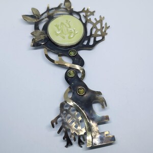 May include: A metal brooch shaped like a key with a dark oxidized finish and polished gold-tone accents. The head features a circular pale green inlay with a raised frog relief, surrounded by branch-like cutouts and three small green gemstones.