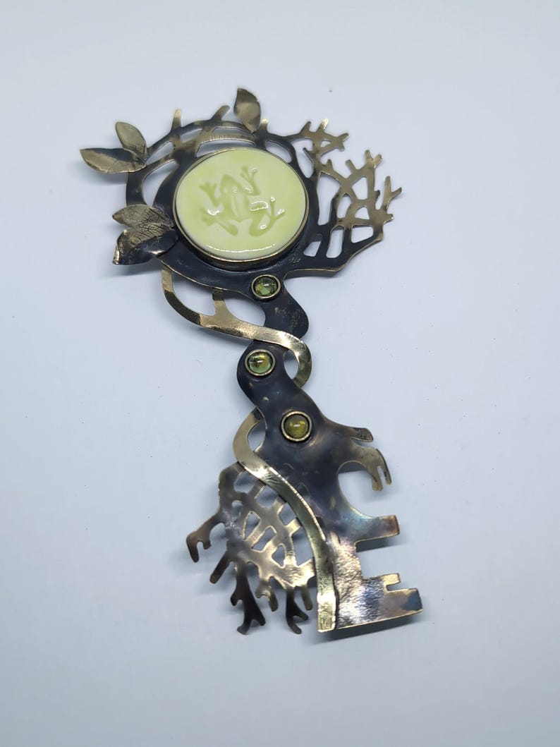 May include: A metal brooch shaped like a key with a dark oxidized finish and polished gold-tone accents. The head features a circular pale green inlay with a raised frog relief, surrounded by branch-like cutouts and three small green gemstones.