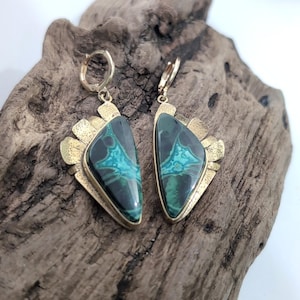 May include: A pair of drop earrings featuring triangular stones with dark green and bright turquoise marbled patterns. The stones are set in textured gold-tone metal frames with hinged hoop closures at the top, showcasing a distinct geometric design.