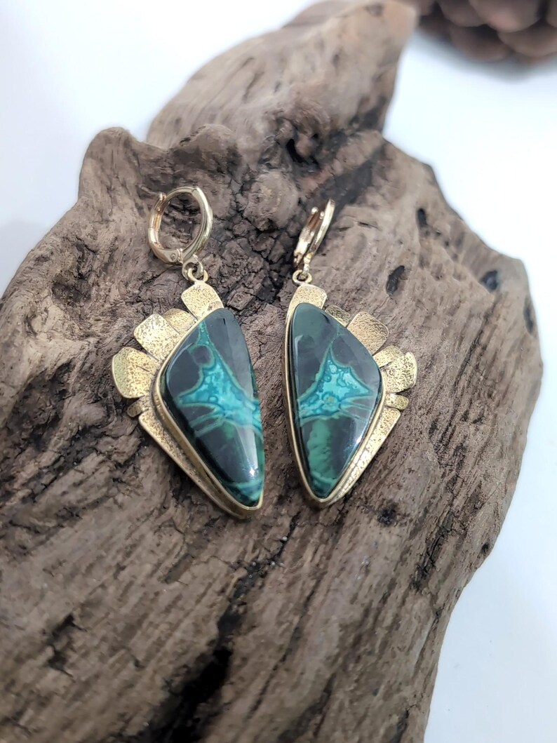 May include: A pair of drop earrings featuring triangular stones with dark green and bright turquoise marbled patterns. The stones are set in textured gold-tone metal frames with hinged hoop closures at the top, showcasing a distinct geometric design.