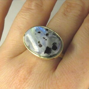 May include: A gold ring featuring an oval-shaped, iridescent gemstone with black inclusions. The stone displays a mix of white, blue, and black colors. The ring is set in a gold band.