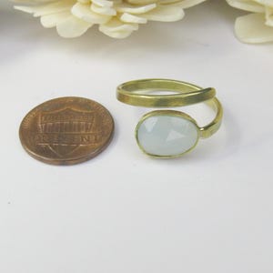 May include: A gold-toned bypass ring featuring a single oval, faceted, pale blue stone. The ring is positioned next to a United States one-cent coin for scale, which has a diameter of approximately 0.75 inches, providing a clear size reference.