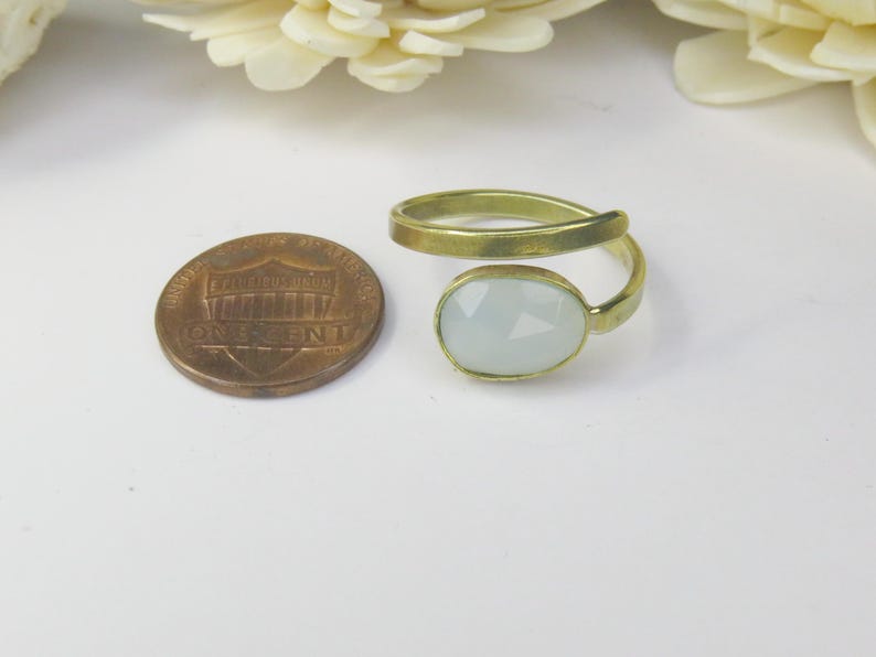 May include: A gold-toned bypass ring featuring a single oval, faceted, pale blue stone. The ring is positioned next to a United States one-cent coin for scale, which has a diameter of approximately 0.75 inches, providing a clear size reference.