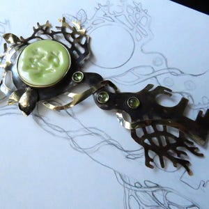 May include: A decorative metal brooch featuring a central pale green circular cabochon surrounded by an intricate, branch-like frame. The piece includes three small, round, light green faceted stones set into the dark, oxidized metal structure.