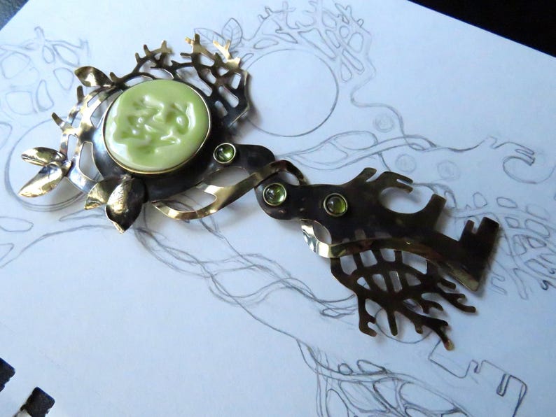 May include: A decorative metal brooch featuring a central pale green circular cabochon surrounded by an intricate, branch-like frame. The piece includes three small, round, light green faceted stones set into the dark, oxidized metal structure.