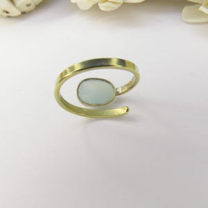 May include: A gold-toned open band ring featuring a single, smooth, oval-shaped pale blue stone set in a bezel. The ring has a bypass design where the ends do not meet, creating a gap between the band and the central stone setting.