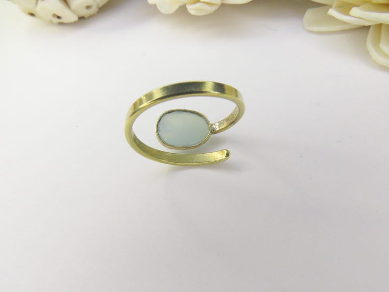May include: A gold-toned open band ring featuring a single, smooth, oval-shaped pale blue stone set in a bezel. The ring has a bypass design where the ends do not meet, creating a gap between the band and the central stone setting.