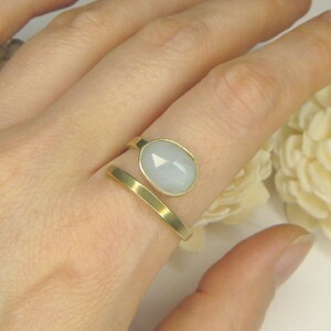 May include: A gold-toned open ring featuring a smooth, flat band and a single oval-shaped, faceted, pale blue-grey stone set in a gold-toned bezel. The ring is designed with a bypass style, leaving a gap between the stone and the band end.