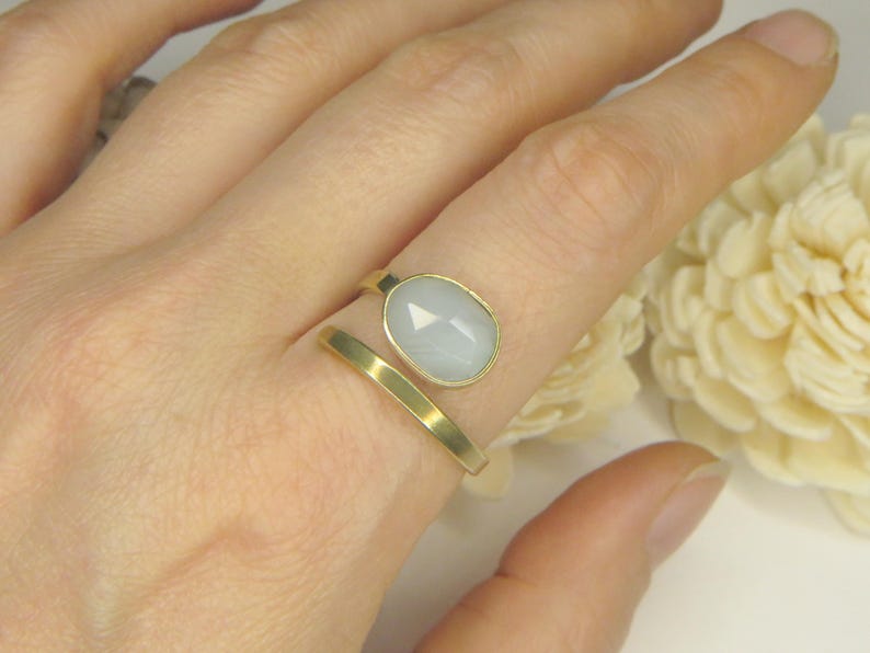 May include: A gold-toned open ring featuring a smooth, flat band and a single oval-shaped, faceted, pale blue-grey stone set in a gold-toned bezel. The ring is designed with a bypass style, leaving a gap between the stone and the band end.
