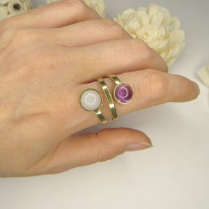 May include: A gold-toned bypass ring featuring two circular settings. One setting contains a smooth, opaque white stone, while the other holds a translucent, deep purple stone. The band has a flat, polished finish and wraps around the finger.