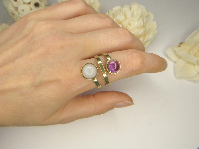 May include: A gold-toned bypass ring featuring two circular settings. One setting contains a smooth, opaque white stone, while the other holds a translucent, deep purple stone. The band has a flat, polished finish and wraps around the finger.