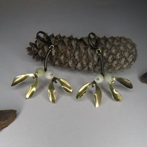 May include: A pair of gold mistletoe earrings with white pearl accents. The earrings are hanging from a brown pine cone.